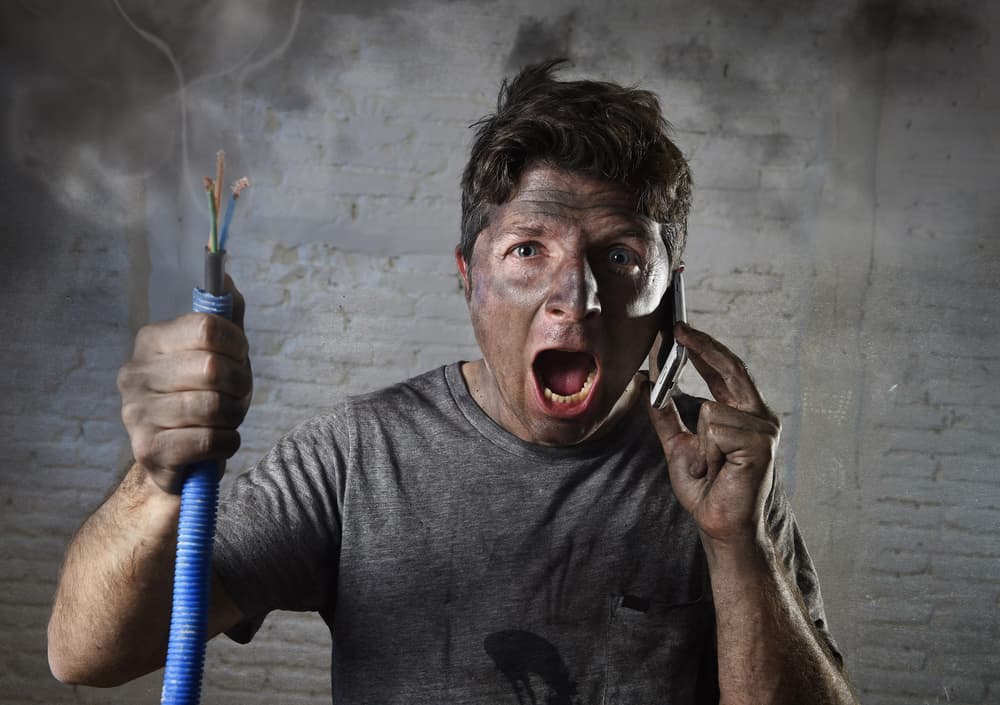 Young man holding electrical cable smoking after electrical accident with dirty burnt face in funny desperate expression calling for help.