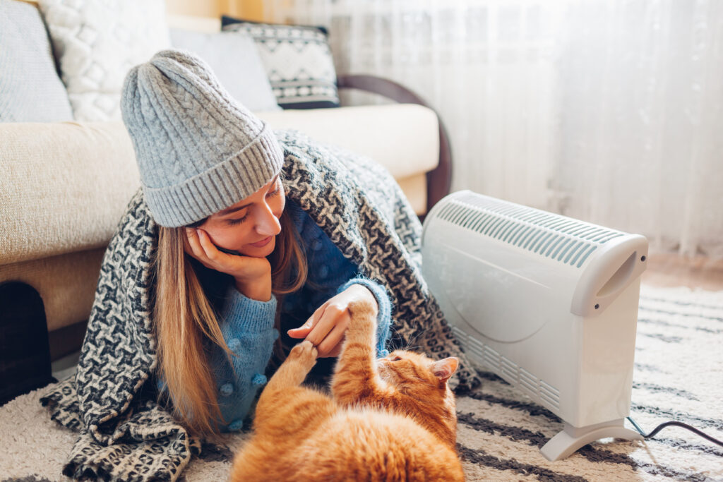 Using heater at home in cold winter. Woman playing with cat by device wearing sweater and hat under blanket. Heating season.